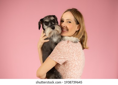 Cute Young Woman Hugs Her Puppy Schnauzer Dog. Love Between Owner And Dog Isolated Over Pink Background. Studio Portrait.