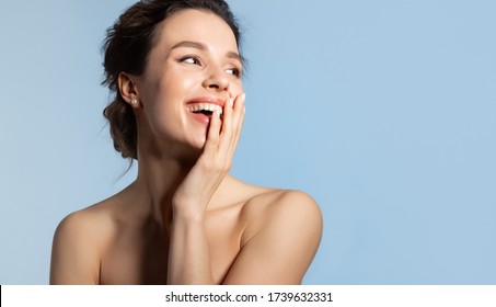 Cute young woman laughing loudly covering mouth with hand looking aside studio portrait on blue copy space. Beautiful girl with naked shoulder, shiny glowing perfect skin isolated headshot