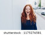 Cute young woman with a lovely sense of humour standing leaning against a white exterior wall with copy space in an urban street laughing at the camera