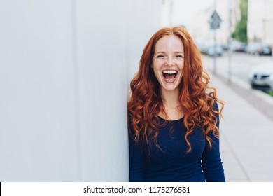 Cute young woman with a lovely sense of humour standing leaning against a white exterior wall with copy space in an urban street laughing at the camera - Powered by Shutterstock