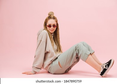 Cute young woman in narrow sunglasses with stylish blond afro braids sitting sideways on the floor over pink background. Wearing loose grey clothes and sneakers.