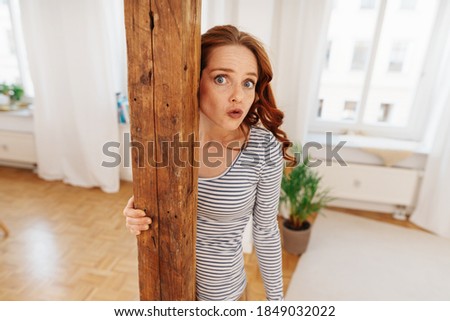Cute young woman pulling a quirky face pursing her lips with wide eyes as she peers around a wooden interior pillar