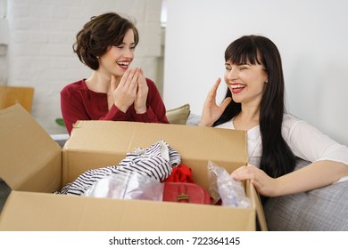 Cute Young Woman Showing Her Humour For A Friends New Garments As They Unpack Them From A Box Laughing Hysterically