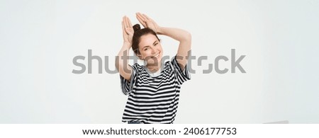 Cute young woman shows animal ears gesture above head, smiling and looking happy, posing in casual clothes over white background.