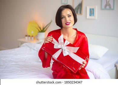 Cute Young Woman Sitting On The Bed With Red Gift. Sexy Woman In Red Silk Dressing Gown With Christmas, Valentine Day Or Birthday Gift