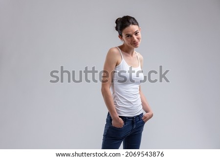 Cute young woman with a sweet smile and dimples standing looking at the camera with hands in the pockets of her jeans over a grey studio background