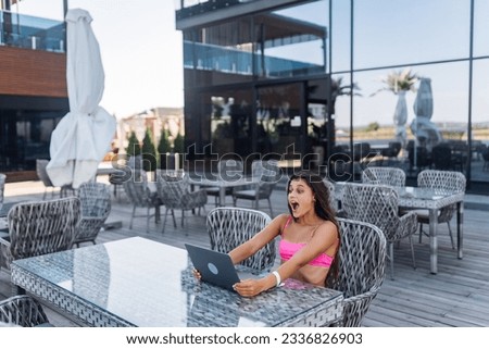 Cute young woman in swimsuit is amazed with a laptop sitting on terrace on the beach