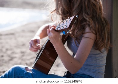 Cute Young Woman Wearing Long Blond Hair Playing  Guitar Sitting On The Beach