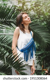 A Cute Young Woman In A White Blouse, Skirt And Blue Leather Fringe Bag Stands Among Tropical Plants.