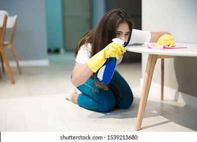 cute young woman in yellow gloves with detergent spray in her hand wiping dust off stain from the kitchen chair with a rag thoroughly