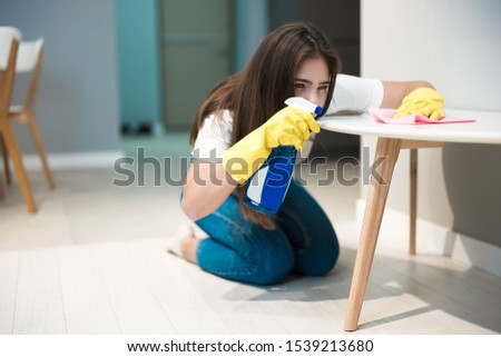 cute young woman in yellow gloves with detergent spray in her hand wiping dust off stain from the kitchen chair with a rag thoroughly
