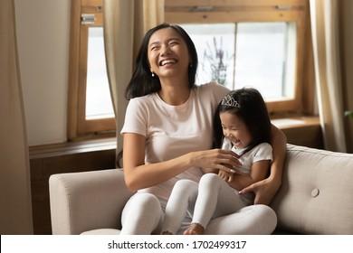 Cutie little daughter seated on mommy lap on couch laughing enjoy funny active time with loving parent. Asian excited young mother tickling adorable preschooler kid girl feels happy be mother concept