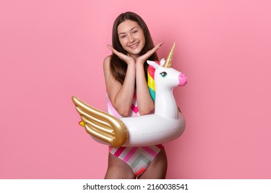 cutie smiling tanned teen girl with inflatable unicorn ring hold hands near face looking at the camera, dressed in a colourful striped one-piece swimsuit isolated on pink background, Summer Vacation