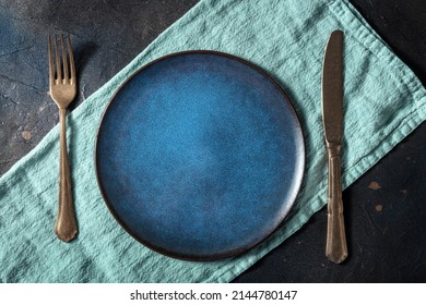 Cutlery With A Blue Plate. A Fork, A Knife, And A Dish On A Teal Napkin, On A Black Slate Background, Overhead Flat Lay Shot