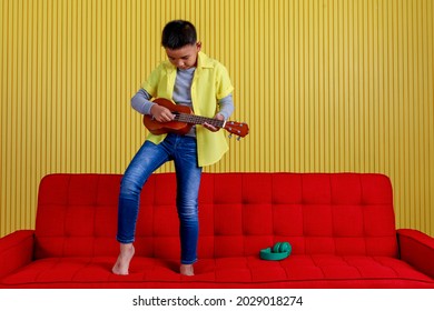 Cutout Portrait Of Young Asian Boy On Grey Sweater, Yellow Shirt, And Blue Jeans Enjoy Acting Music Show As Professional Singer, Smart Musician By Confidently Playing Small Guitar Near Striped Wall