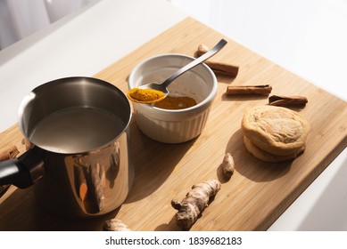 Cutting board with curcuma powder in white bowl,  pot with almond milk, cinnamon sticks and row turmeric roots on wooden background. Preparation steps of homemade curcuma latte.