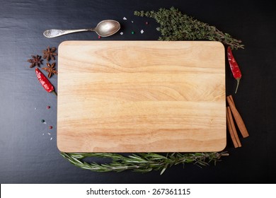 Cutting board with rosemary, thyme, colored and chili pepper, old spoon, cinnamon, cardamom and sea salt on a black background.
