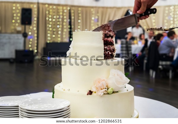 Cutting the first tier of white mastic wedding cake\
in banquet hall background. Front view at table with a beautiful\
wedding cake and side plates for serving guests. Dividing wedding\
cake into parts