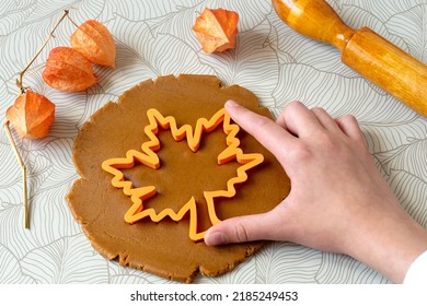 Cutting out autumn gingerbread in form of maple leaf from dark honey dough with spices. Dough, rolling pin, physalis lanterns and cookie cutters on table