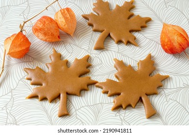 Cutting out autumn gingerbread in form of maple leaf from dark honey dough with spices. Dough, physalis lanterns and cut out raw cookies on table