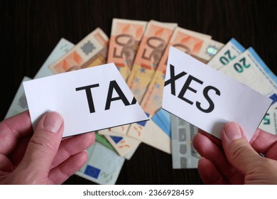 the cutting paper with inscription taxes lies on top of a large pile of euro money on a dark wooden table, top view, tax reduction concept - Shutterstock ID 2366928459