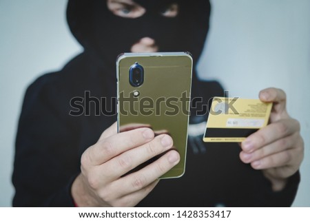 Cyber criminal in balaclava enters the information of a personal bank account. Credit card fraudulent scheme. Stealing cyber money using mobile. New ways of fraudulent transactions via online banking.