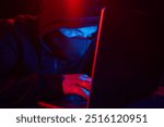 Cyber hacker in hood behind laptop on dark background with red blue light, anonymous spy. digital cyber security concept