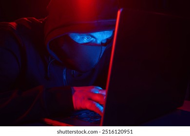 Cyber hacker in hood behind laptop on dark background with red blue light, anonymous spy. digital cyber security concept