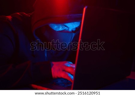 Cyber hacker in hood behind laptop on dark background with red blue light, anonymous spy. digital cyber security concept