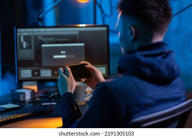 cybercrime, hacking and technology concept - close up of male hacker with smartphone using computer virus program for cyber attack in dark room - Shutterstock ID 2191305103