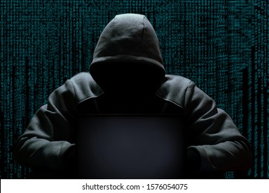 A cybercriminal in a hood without a face sits in the dark behind a laptop, against the backdrop of a binary code