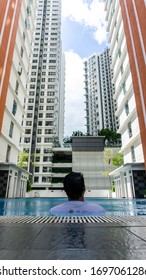 Cyberjaya, Malaysia - March 1, 2020: Back View Portrait Of Young Malay Man Leaning At The Side Of The Swimming Pool Looking At The Tall Building In The City. Luxury Lifestyle Concept. 