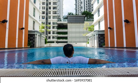 Cyberjaya, Malaysia - March 10, 2020: Back View Portrait Of Young Malay Man Leaning At The Side Of The Swimming Pool Looking At The Tall Building In The City. Luxury Lifestyle Concept. 