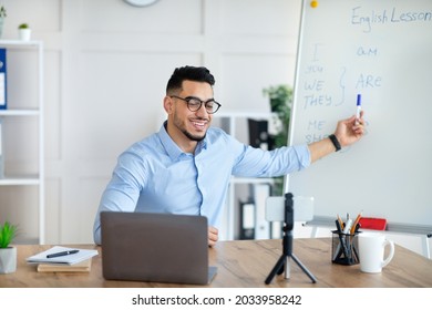 Cyberlearning, remote education concept. Happy Arab male teacher giving online English lesson, using smartphone and laptop, pointing at blackboard with grammar rules at home office - Shutterstock ID 2033958242