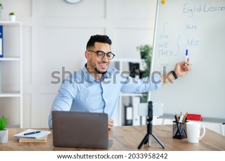 Cyberlearning, remote education concept. Happy Arab male teacher giving online English lesson, using smartphone and laptop, pointing at blackboard with grammar rules at home office