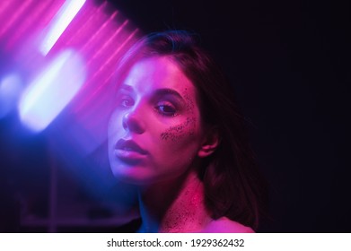 Cyberpunk Photo Of Attractive Woman In Colored Light On Dark Background With Glitter On Face Posing At Camera In Blue And Purple Light With Glare.