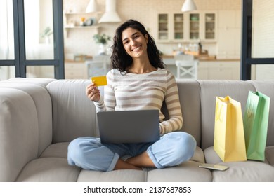 Cybershopping Concept. Smiling Young Lady Sitting Cross Legged On Couch, Surrounded By Gift Bags, Using Laptop And Credit Card, Buying Goods In Online Store At Home, Full Length