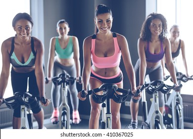 Cycling beauties. Young beautiful women with perfect bodies in sportswear looking at camera with smile while cycling at gym