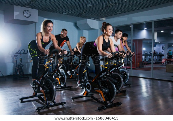 Cycling class in fitness club, group of fit\
people spinning on cardio machine. Man and women do sports\
exercises at gym fat burning class. Active lifestyle, health care\
and body training\
concept