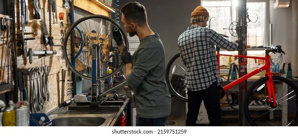 Cycling mechanic checking bicycle wheel spoke with bike spoke wrench while his male colleague checking or fixing bicycle in workshop. Young caucasian servicemen. Bike service, repair and upgrade - Shutterstock ID 2159751259