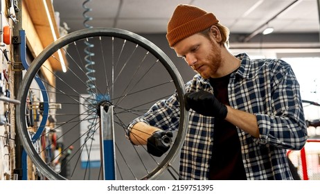 Cycling mechanic checking bicycle wheel spoke with bike spoke wrench in modern workshop. Young focused caucasian redhead man. Bike service, repair and upgrade. Garage interior with tools and equipment