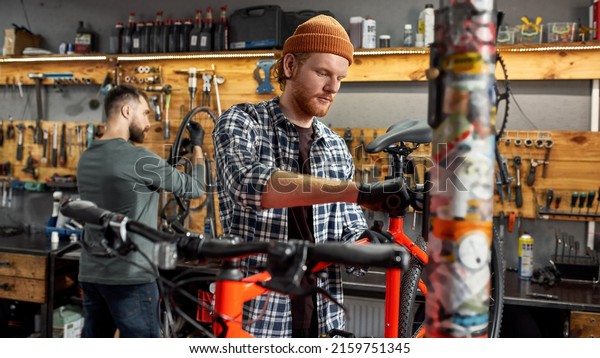 Cycling mechanic fixing bicycle seat while his\
male colleague checking bicycle wheel spoke with bike spoke key on\
background in workshop. Young caucasian repairmen. Bike service,\
repair and upgrade