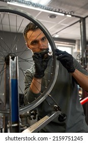 Cycling mechanic or technician checking bicycle wheel spoke with bike spoke key in modern blurred workshop. Young concentrated caucasian bearded man. Bike service, repair and upgrade