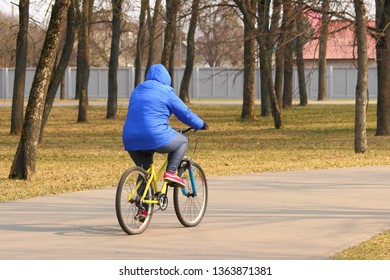 Cycling Outdoor – An Older Woman In A Blue Jacket With A Hood Riding A Mountain Bicycle On Asphalt Alley In Park On A Spring Day, Rear View