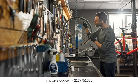 Cycling repairman checking bicycle wheel spoke with bike spoke key in modern workshop. Young caucasian bearded man. Bike service, repair and upgrade. Garage interior with tools and equipment