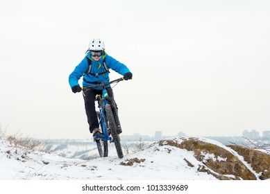 Cyclist In Blue Riding The Mountain Bike On The Rocky Winter Hill Covered With Snow. Extreme Sport And Enduro Biking Concept.