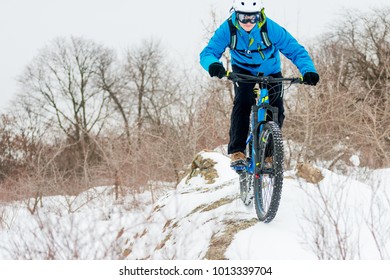 Cyclist In Blue Riding The Mountain Bike On The Rocky Winter Hill Covered With Snow. Extreme Sport And Enduro Biking Concept.