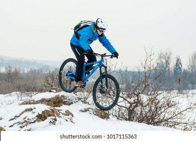 Cyclist In Blue Riding The Mountain Bike On The Rocky Winter Hill Covered With Snow. Extreme Sport And Enduro Biking Concept.