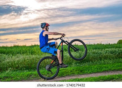 A Cyclist In A Helmet Rides A Bicycle On The Rear Wheel During Sunset. Wheelie.