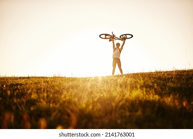 The cyclist is holding his bicycle over himself on the background of red sunset. Biker with bicycle on the field during sunrise. High quality photo